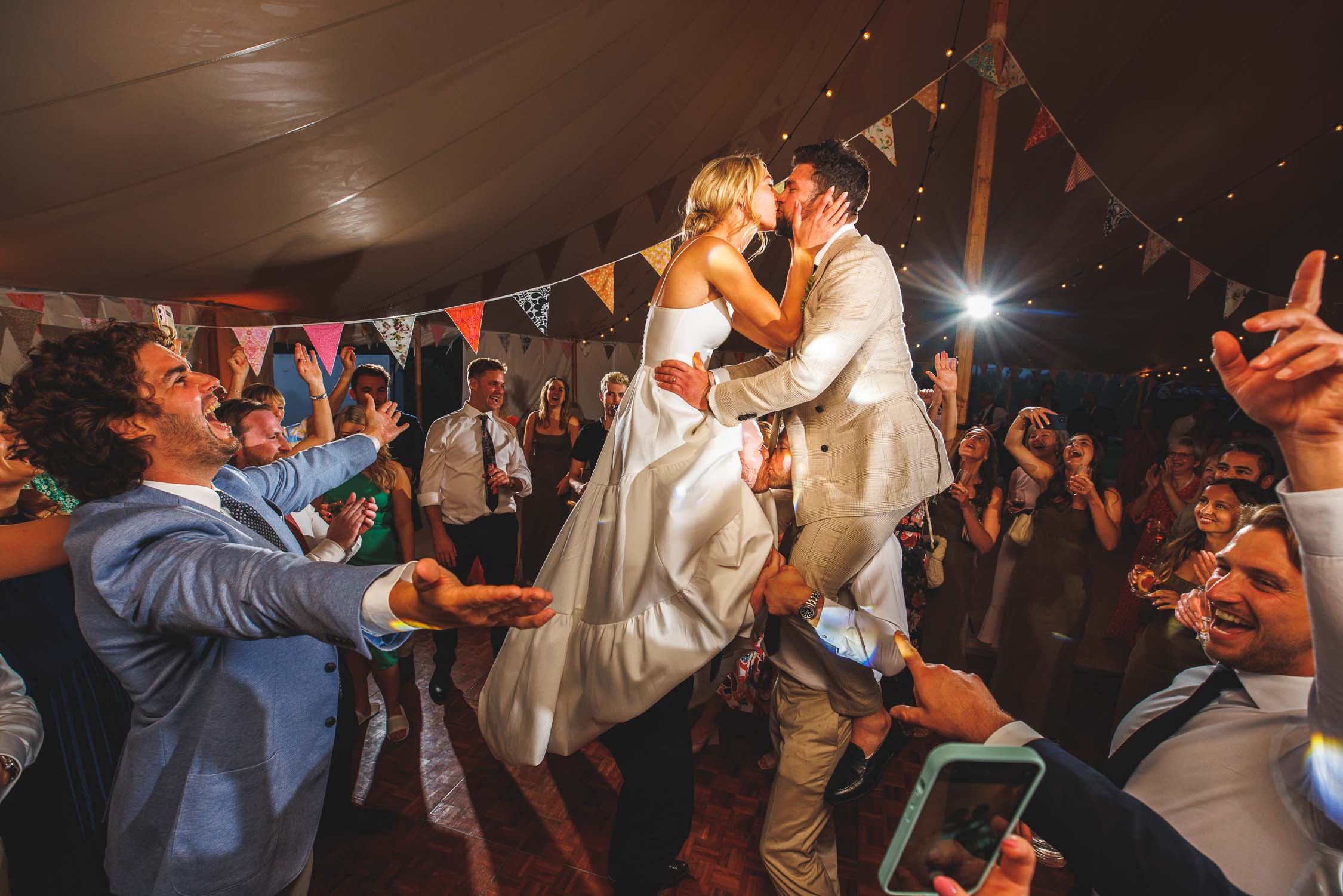 The bride and groom kiss while raised up on shoulders surrounded by their wedding guests. Taken at a wedding at Pauntley Court in Gloucestershire.